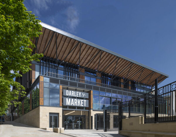 The modern entrance to Darley Street Market, featuring a timber and steel canopy roof, floor-to-ceiling glazing, and ornate black iron gates against a bright blue sky.